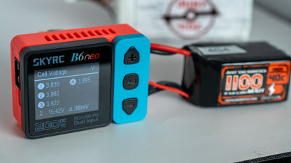 SKYRC B6neo battery monitor with a battery pack in the background on a light surface.